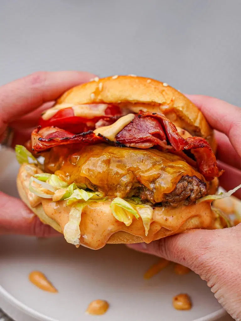 Two hands gripping a beef and bacon burger, ready for a big bite.
