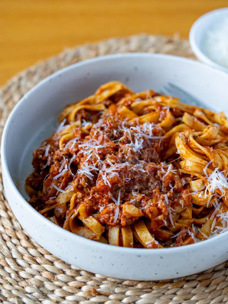 Bowl of fettuccine pasta topped with hidden vegetable Bolognese sauce and freshly grated Parmesan cheese, served on a woven placemat.