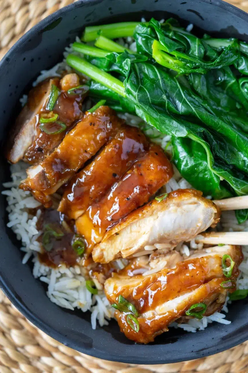 Honey garlic chicken being picked up with chopsticks from a bowl of rice and greens.