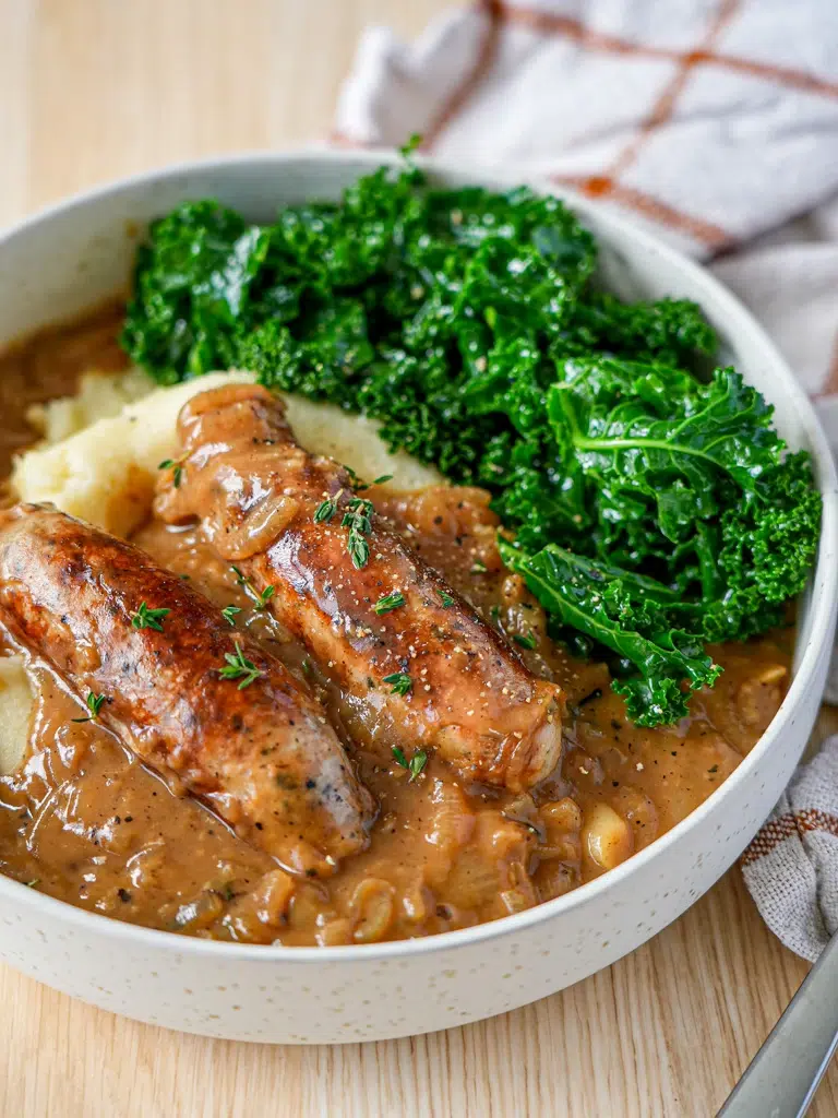 A bowl of Sausages served with homemade onion gravy, garnished with fresh herbs, and accompanied by mashed potatoes