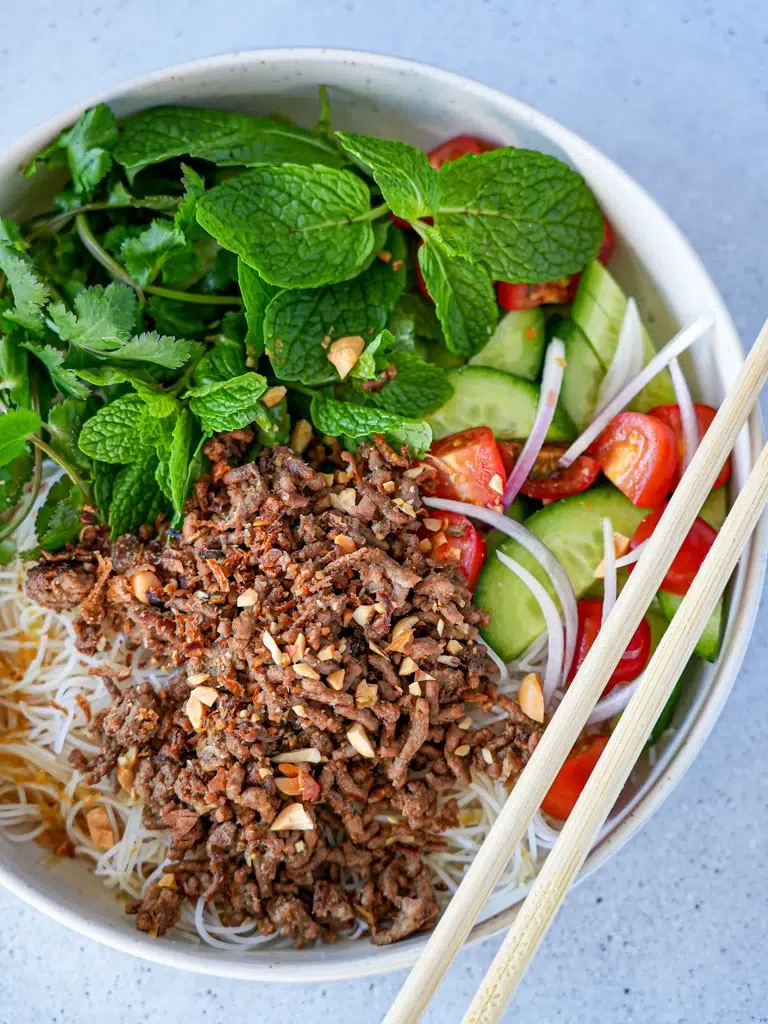 Bowl of Thai beef and noodle salad featuring seasoned ground beef, topped with sliced cucumber, rice noodles, fresh herbs, and onions, with chopsticks on the side